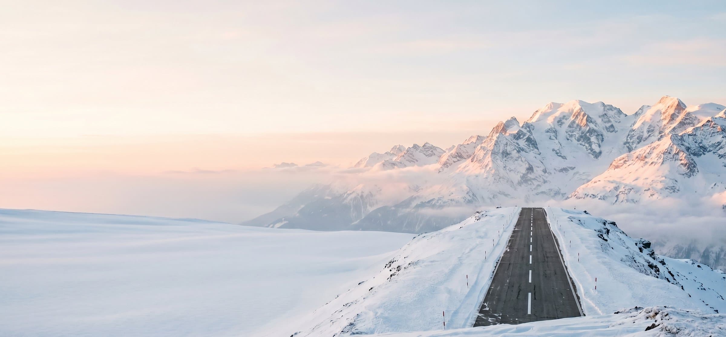 Asphalt runway on a snow-covered alpine ridge at dawn, with snow-capped peaks glowing in warm light